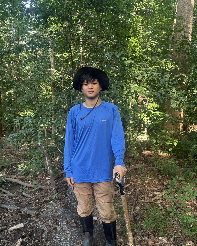 Daniel Tang standing on a woodland trail in a wide-brimmed hat and a
                  blue long-sleeved shirt, trees behind him.