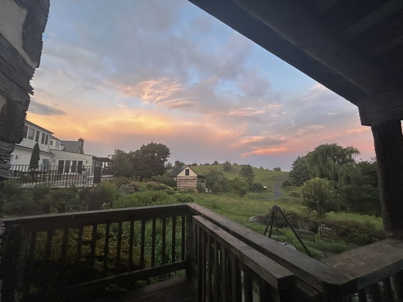 Looking out from a covered porch at sunset over rolling pasture: a
                  farmhouse to the left, a small log cabin below, a willow, and a track
                  running over the hill under pink and orange cloud.